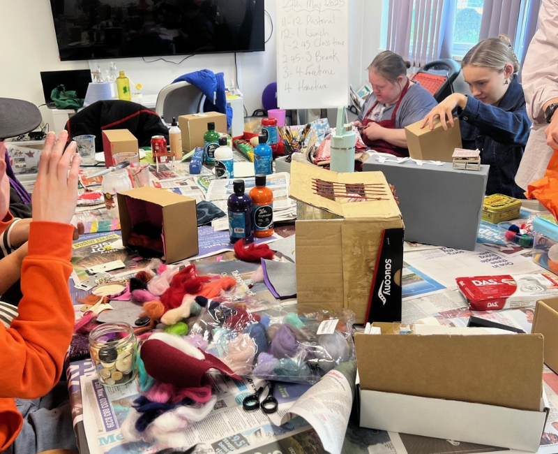 A workshop is in progress, at least four people sit round a table surrounded by arts materials creating their shoeboxes