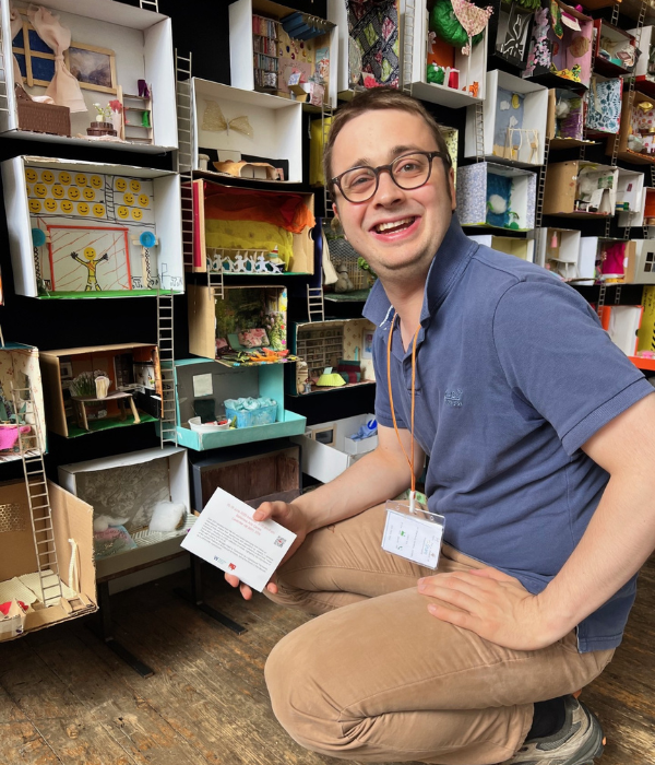 A man kneels in front of the shoebox installation smiling