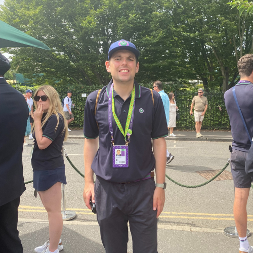 Joe stands in his uniform smiling alongside the queue at Wimbledon