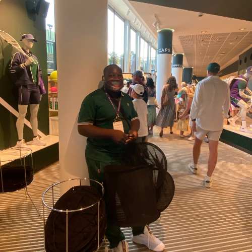 Shammah stands next to a pillar inside the shop he's working in at Wimbledon, he has a pile of bags he's handing out to shoppers.