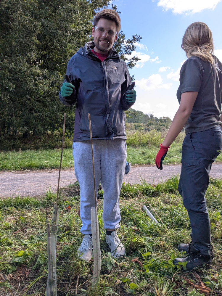 A young man stands smiling at the camera outside in a garden with a garden rake