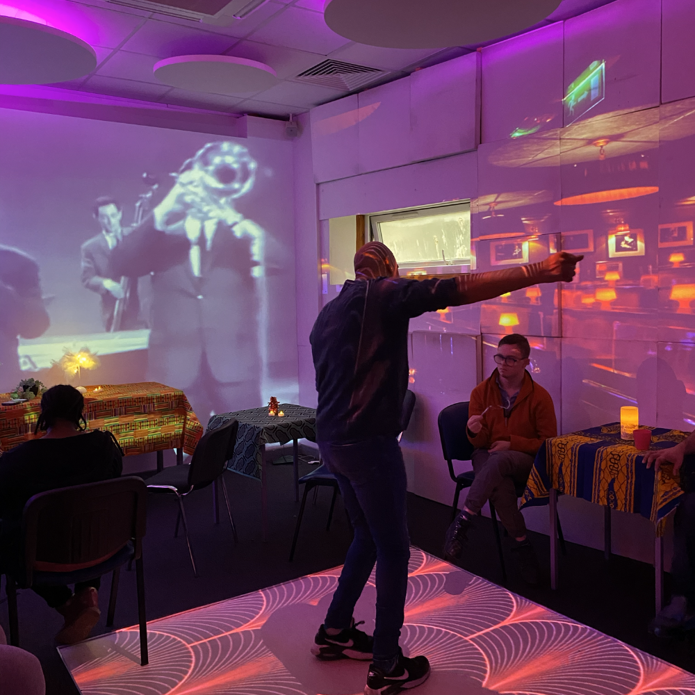 A man dances slowly along to some jazz, behind him on the screen is black and white footage of a jazz musican