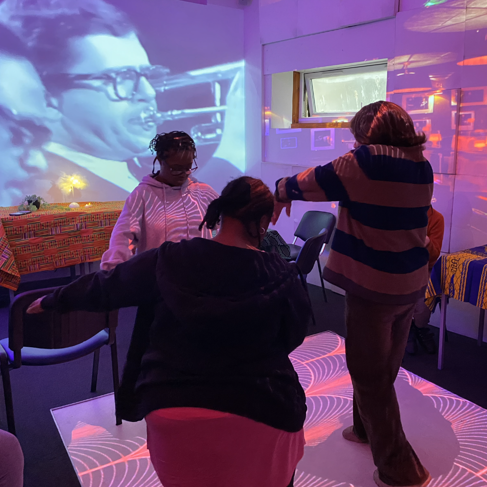 Three women dance on a patterned floor, behind them on a large screen is black and white footage of jazz musicians playing the trumpet.