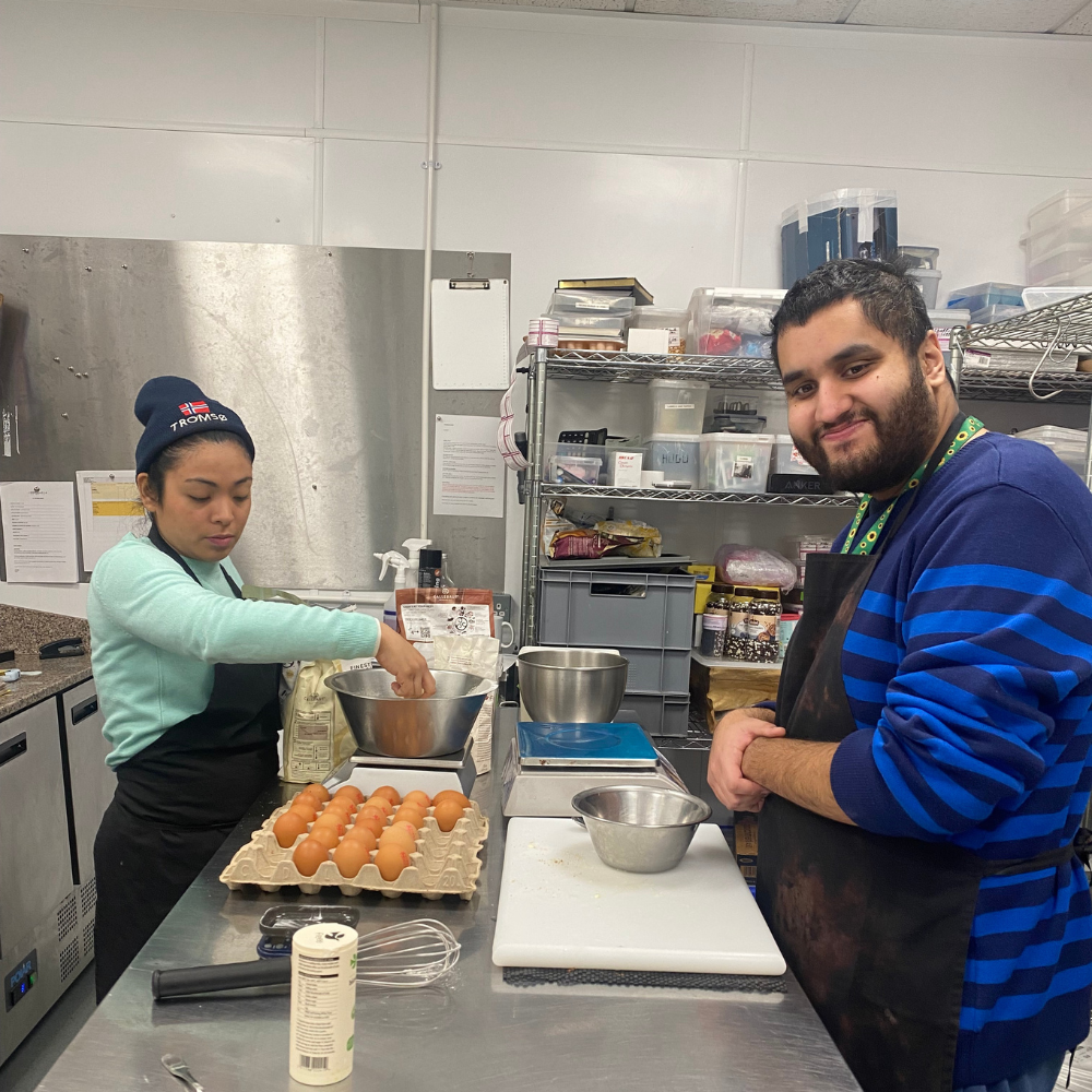 A man, Zain, and a lady, Paula, stand over the prep table in the professional kitchen basting their raw scones with beaten egg ready for backing. They're both wearing aprons and smiling at the camera.