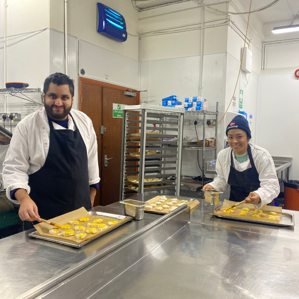 A man, Zain, and a lady, Paula, stand over the prep table in the professional kitchen basting their raw scones with beaten egg ready for backing. They're both wearing aprons and smiling at the camera.