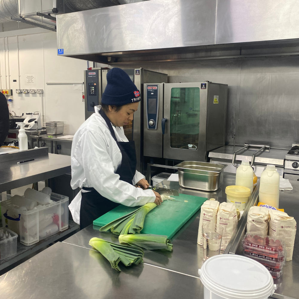 Share student Paula stands at a prep table in a professional kitchen chopping up leeks ready for soup. 