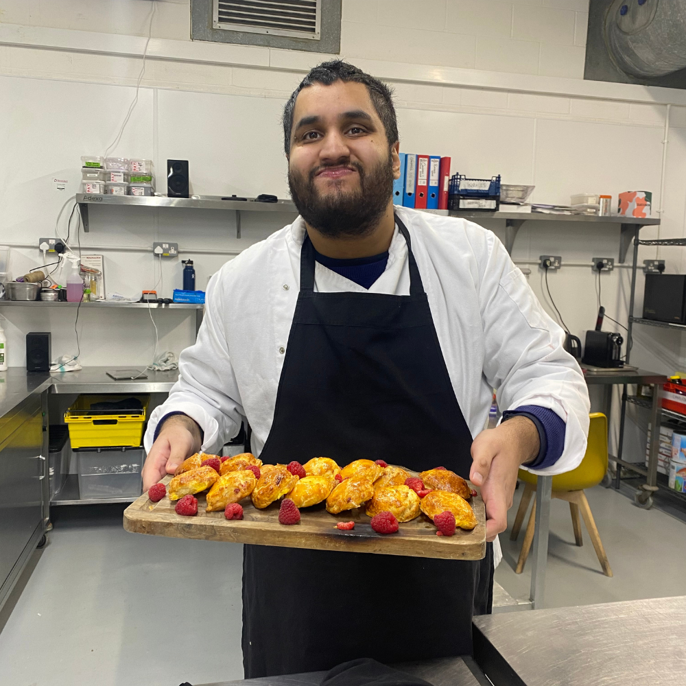A man who is a Share student called Zain stands in a professional kitchen proudly holding a board full of freshly baked scones. He is smiling. 