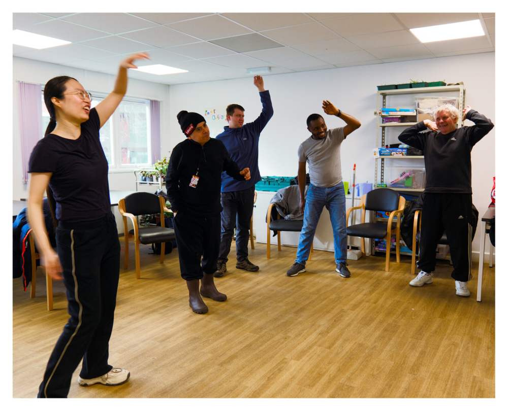 A group of people stand in a classroom with one hand in the air as part of a movement workshop