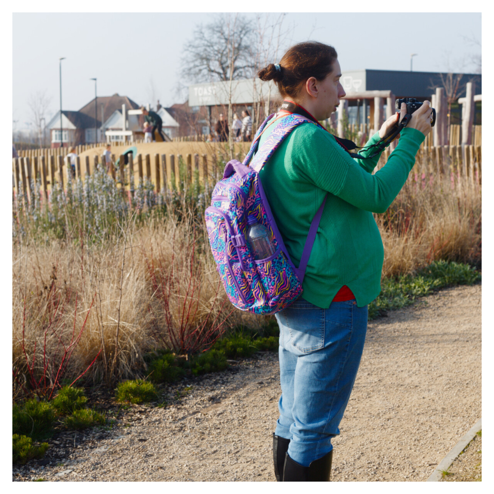 A lady stands on a path in a park taking a photo, she's wearing green top and has a purple backpack