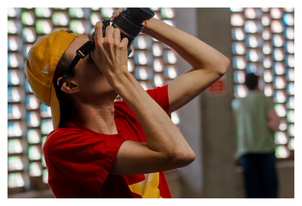A man in a red tshirt and yellow cap aims his camera up high at an angle to capture a good shot