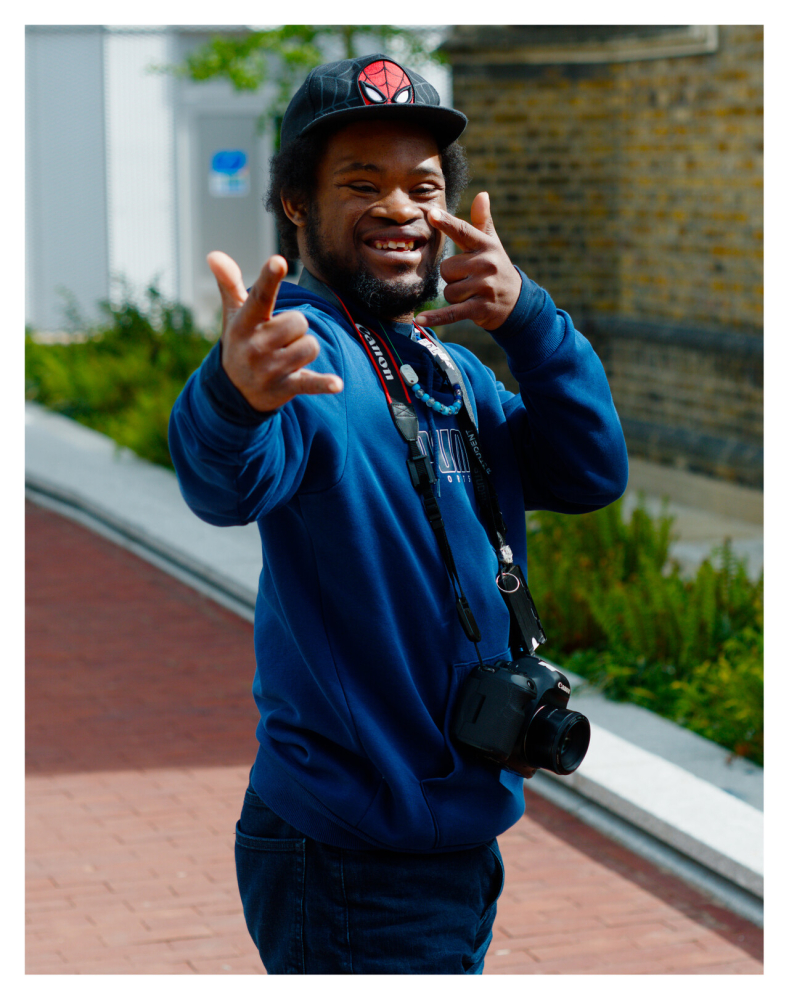 Man in a blue top, cap and with a camera round his neck gives a thumbs up to the camera