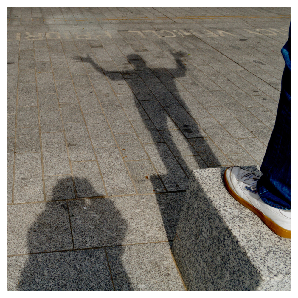 Image of two people's shadows on concrete, one is clearly taking a photo of the other who has hands in the air