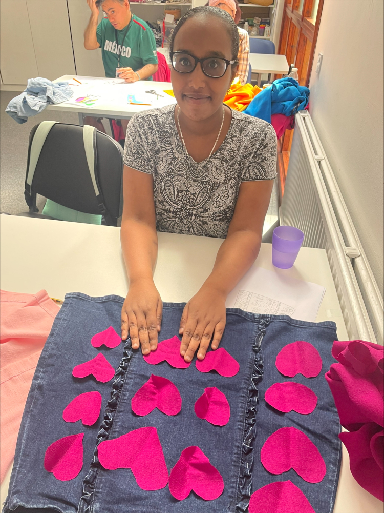 A lady sits smiling above her needlework: pink fabric hearts sewn on a black background fabric