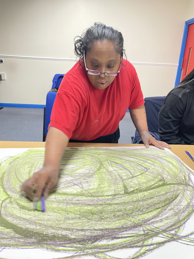 A lady stands over a table making a big arc with her crayons creating a circular artwork in green stripes