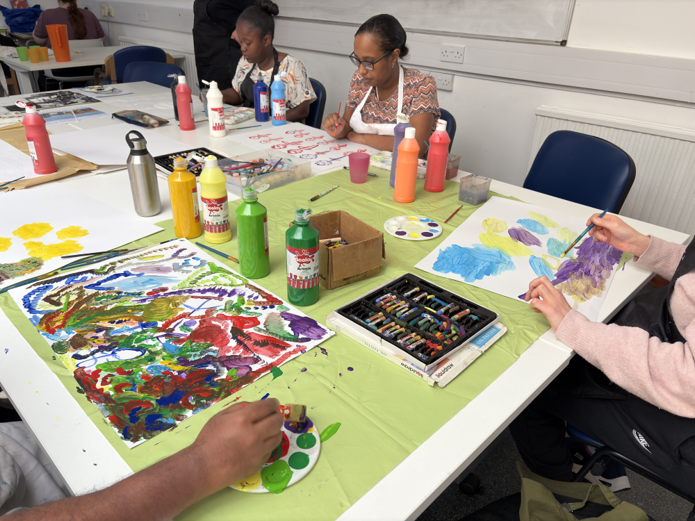 A group of students sit at a large table painting, surrounded by paints and brushes
