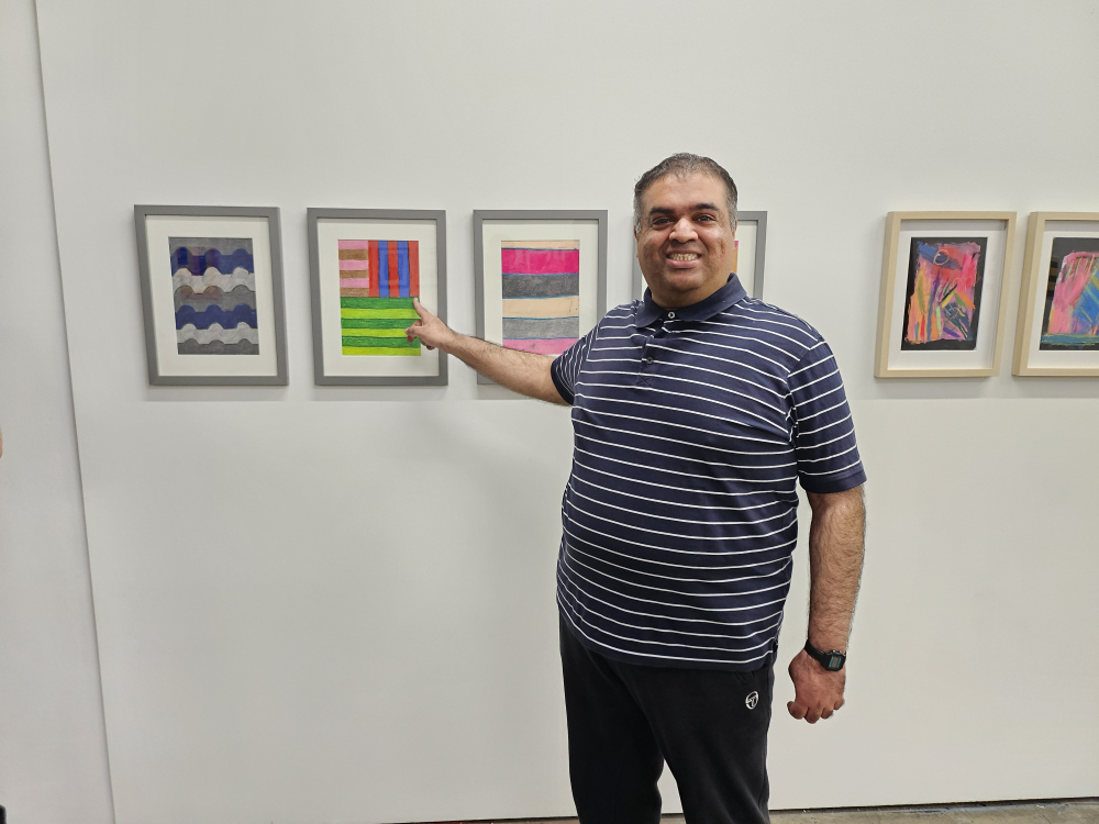 A man stands next to three paintings on the wall pointing at his artwork