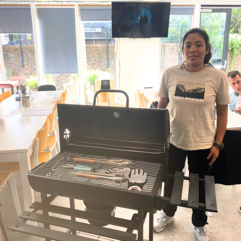 Student Paula stands proudly next to an assembled BBQ inside a cafe, all the BBQ accessories are laid out on it's grill