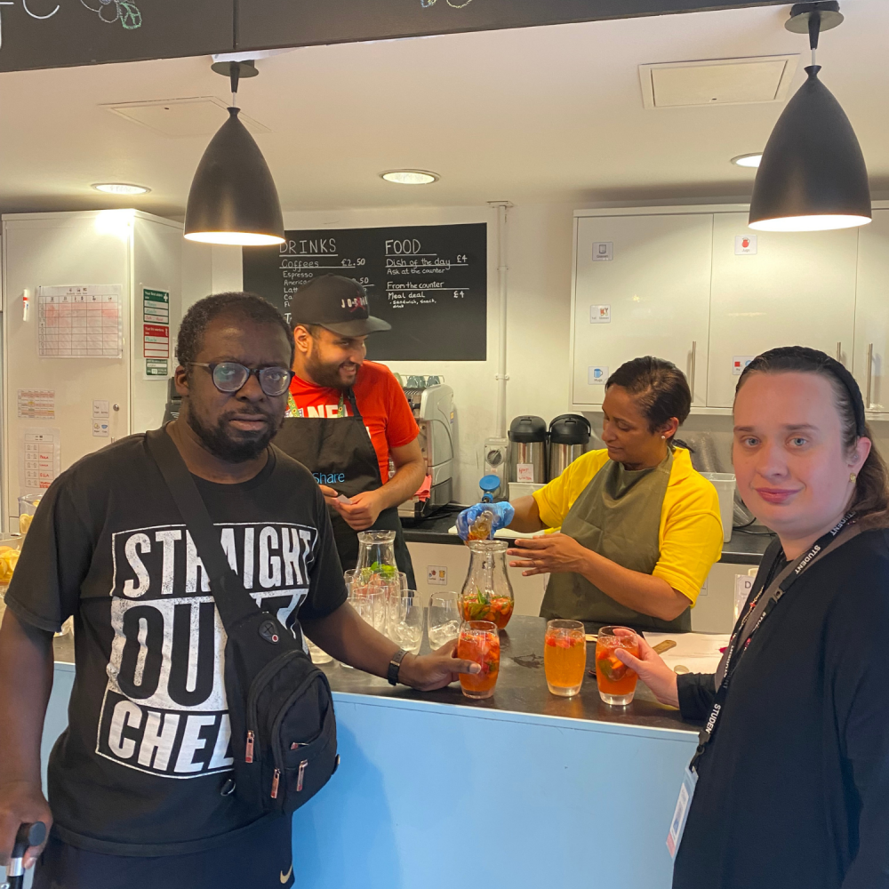 A man and a woman who are student guests at the supper club stand waiting for their mocktails, behind them a student and volunteer are busy pouring drinks at the counter