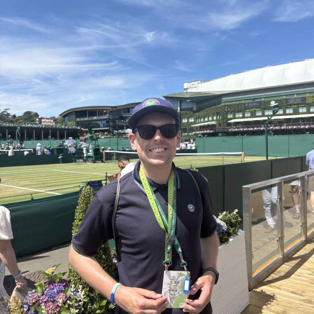 A man stands smiling in his Wimbledon tennis uniform holding his work lanyard