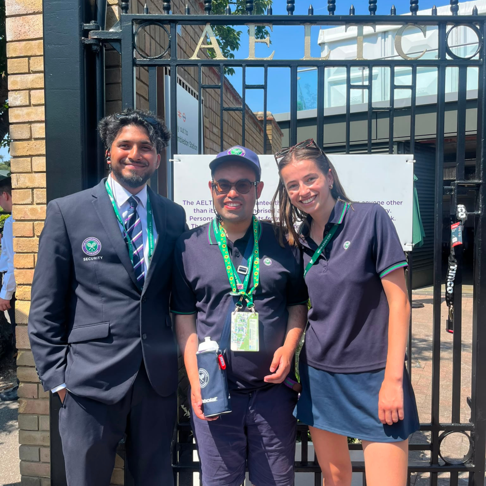 A man in his WImbledon uniform stands proudly outside the Wimbledon gates with his team colleagues