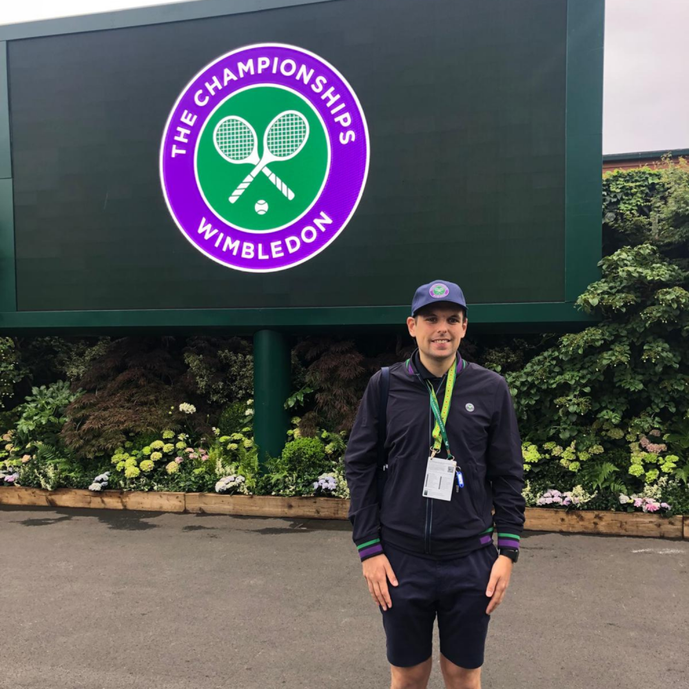 A man stands proudly in his Wimbledon work uniform, behind him a large Wimbledon logo on the wall in the brand colours purple and green