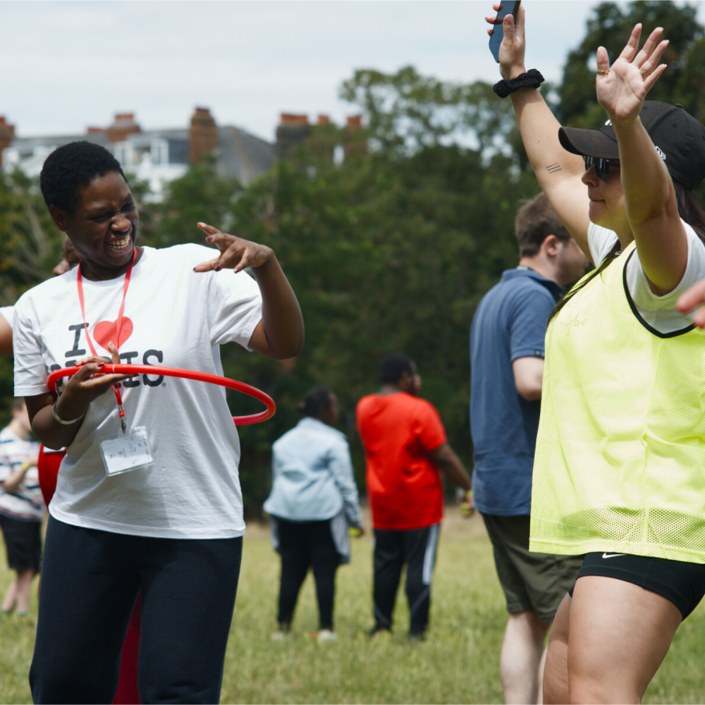 A man is hoola hooping with a red hula hoop and a volunteer in high vis is waving her arms in the air next to him
