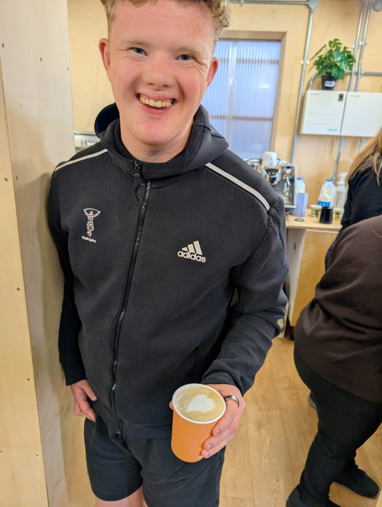 A young man stands smiling showing off a barista style coffee he's just made