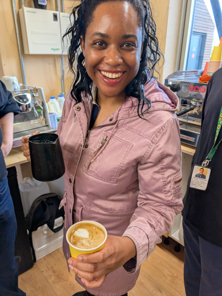 A lady stands smiling proudly holding out a cup of coffee she's just made on a barista machine