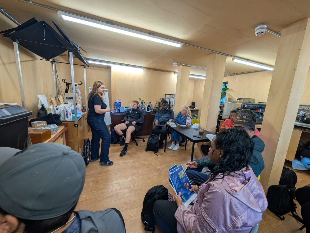 A group sit around listening to a teacher who is telling them about barista technique