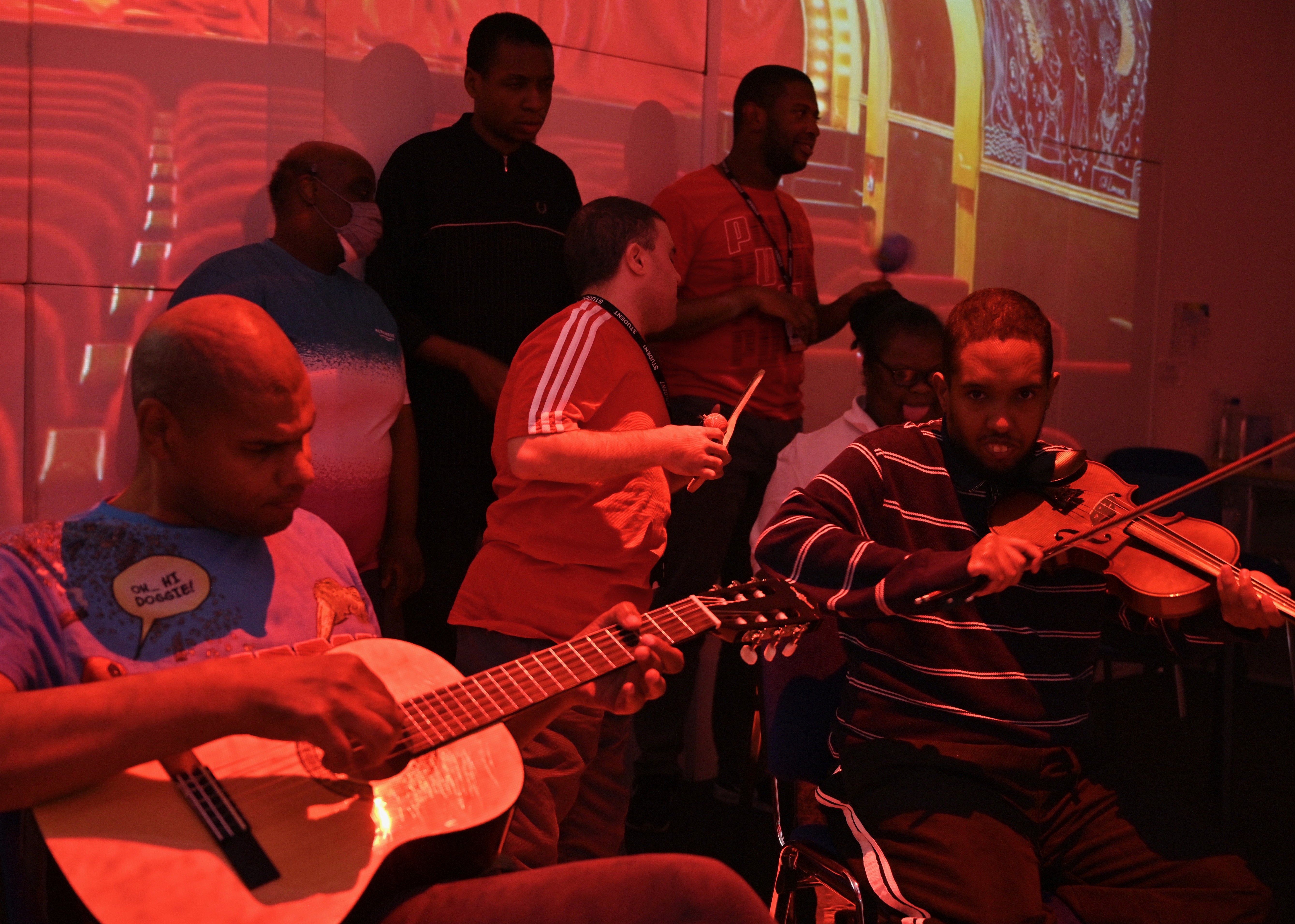 a group of musicians attending a session at Share Community play a mix of instruments including guitar and violin, seated and standing in a room covered in red light, the group are in a room with 4D audio visual equipment that projects coloured light