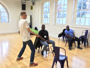 A group of students sit round on chairs in a room with the drama teacher about to crouch down