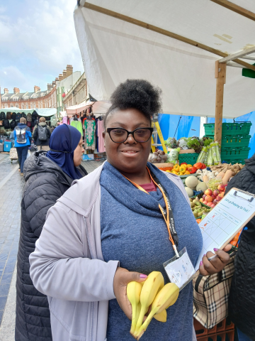 A Share student standing in front of a market stall selling fruit and vegetables. She is holding a banana and a clipboard.