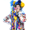 Laughing lady wearing a very extravagant blue and yellow hat with ribbons poses with one arm folded up to her neck and one on her opposite waist