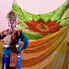Dancer in traditional Indian dress, playing the part of Kubera, stands in front of green orange and red patterned flags