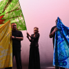 Share students on stage standing in front of green, blue, orange and red patterned flags