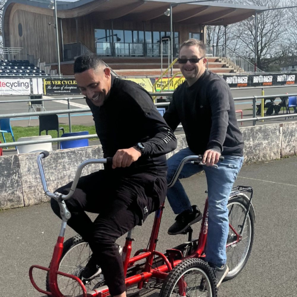 Two men ride on an adult sized tricycle in a velodrome