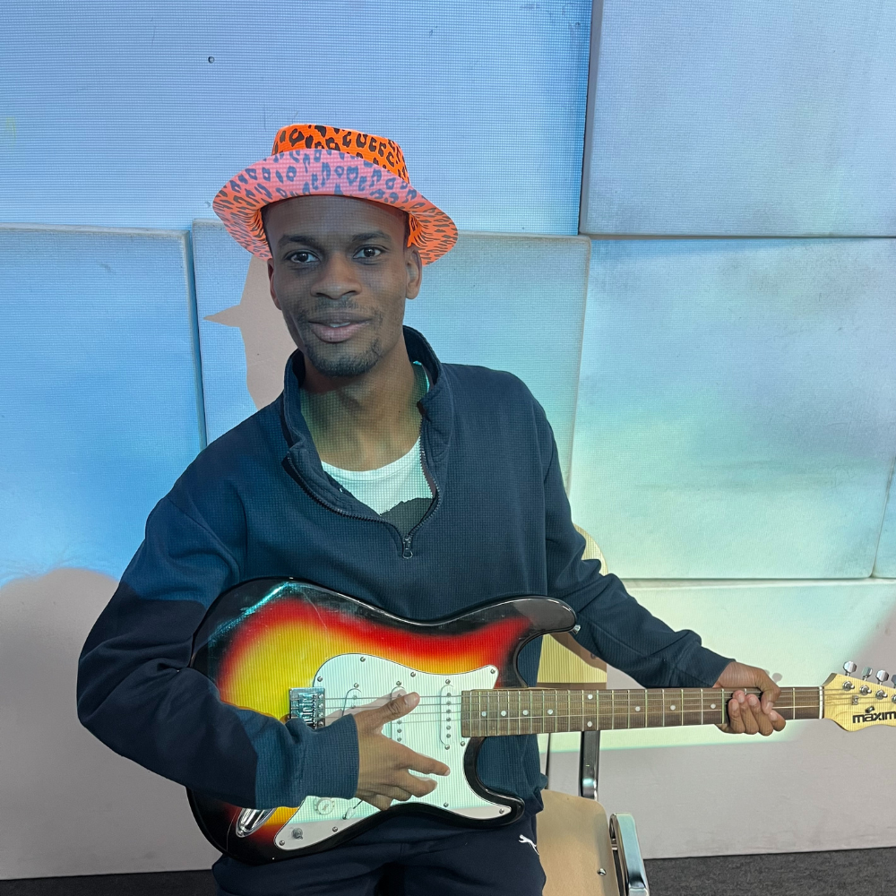 Daniel sits on a chair with a crazy pink hat on and plays electric guitar