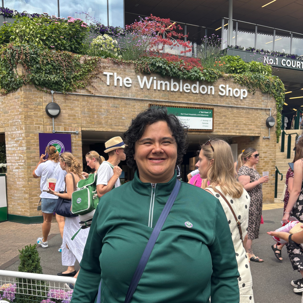 Share student Charlotte stands outside The Wimbledon Shop at the tennis championships. She's in uniform and smiling, having just finished her shift working there