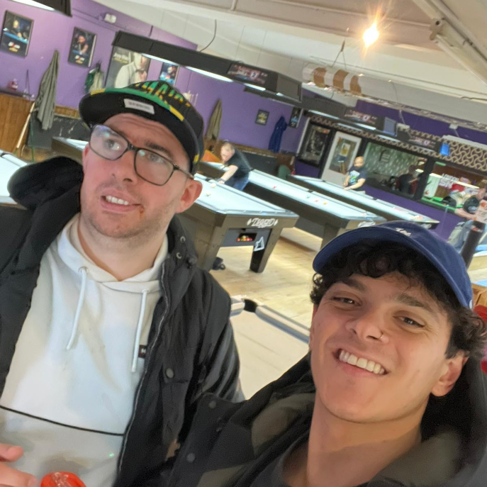 Two men take a selfie, both are wearing baseball caps, behind them is a pool table