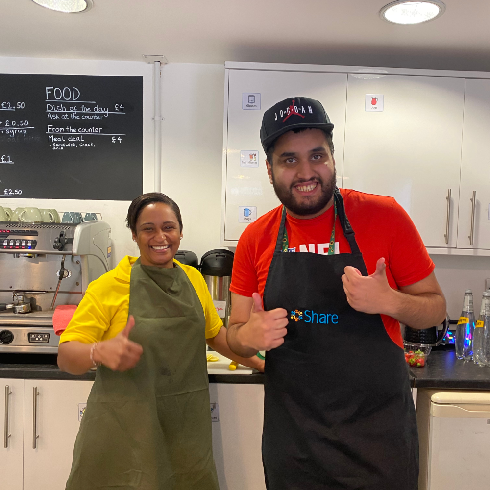 A Share student wearing an apron gives a thumbs up to the camera, he's standing behind a cafe counter with a volunteer lady who also gives a thumbs up and wears an apron, both smiling