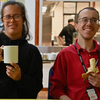Two people sit in a canteen smiling, one is eating a banana, the other holding a mug of tea.