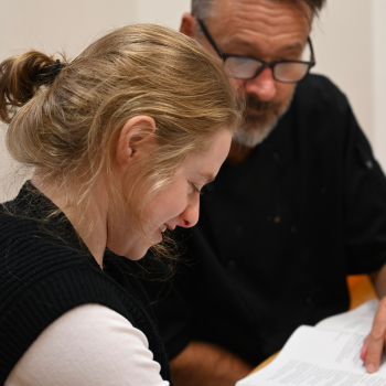 A lady sits working at a desk with a member of staff or tutor helping her.