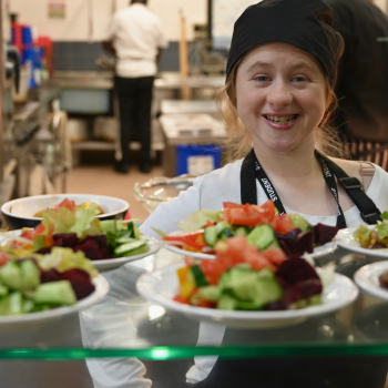 A lady in a catering hat and apron stands smiling proudly over the food she's just prepared.
