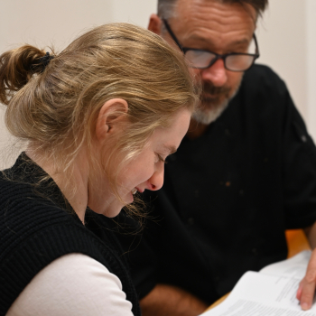 A lady sits looking at some papers on a desk with a member of staff guiding her through