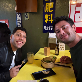 Two men lean in for a selfie over a table in a restaurant, the table has a yellow tablecloth and Chinese writing on the wall suggests they're in an Asian restaurant