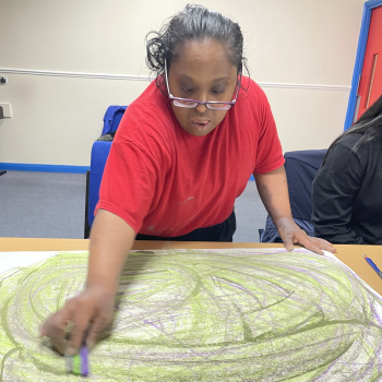 A lady leans over a table where she's drawing a circular design with green and blue arcs, she's wearing a red t-shirt and glasses and is concentrating hard on work.