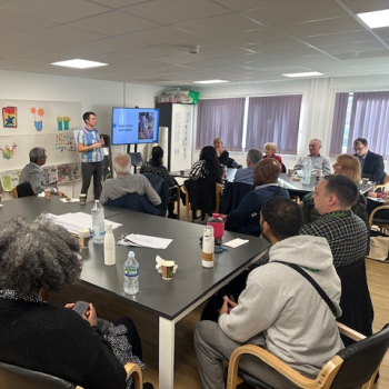 A group of people sit round desks in a seminar, listening to a lady standing at the front talking