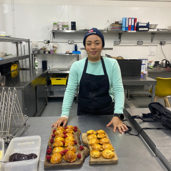 A lady wearing a beanie hat and an apron stands proudly with her batch of baked scones that are on the prep table in front of her. She's in a professional kitchen and is smiling. 