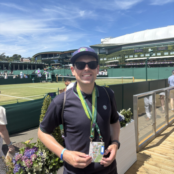 A student stands in Wimbledon work clothes outside a court, wearing a baseball cap and sunglasses. He's smiling and holding his official work lanyard.