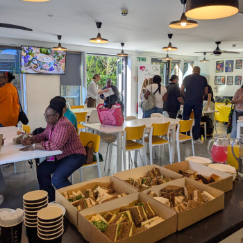 An event with tables where people are talking about services available, in the foreground some cakes and refreshments at the cafe counter